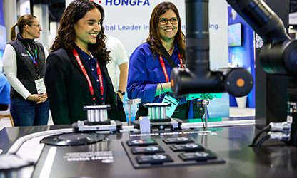 Two women stand beside a machine at an exhibition, engaged in conversation and examining the equipment