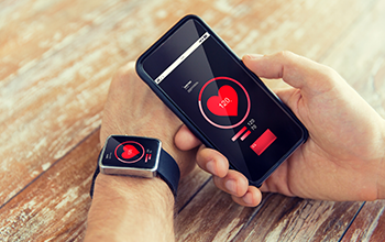 Person checking heart rate on a smartwatch while holding a smartphone displaying the same heart rate data, both devices showing 130 bpm on wooden table.