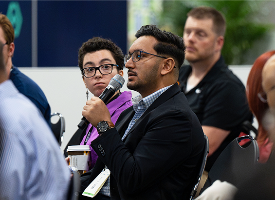 A conference attendee surrounded by other attendees speaks at the audience holding a microphone 