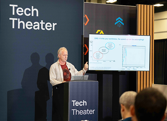 A male conference speaker in a podium pointing to graphics on a board